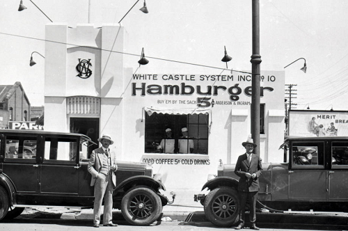 two cars with men standing in near the hoods in front of hamburger shop