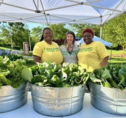 three women stand behind metal pails of lettuce
